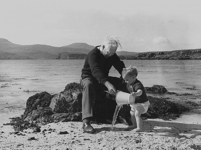 Black and white photograph of a small child playing on a beach with an elderly gentleman