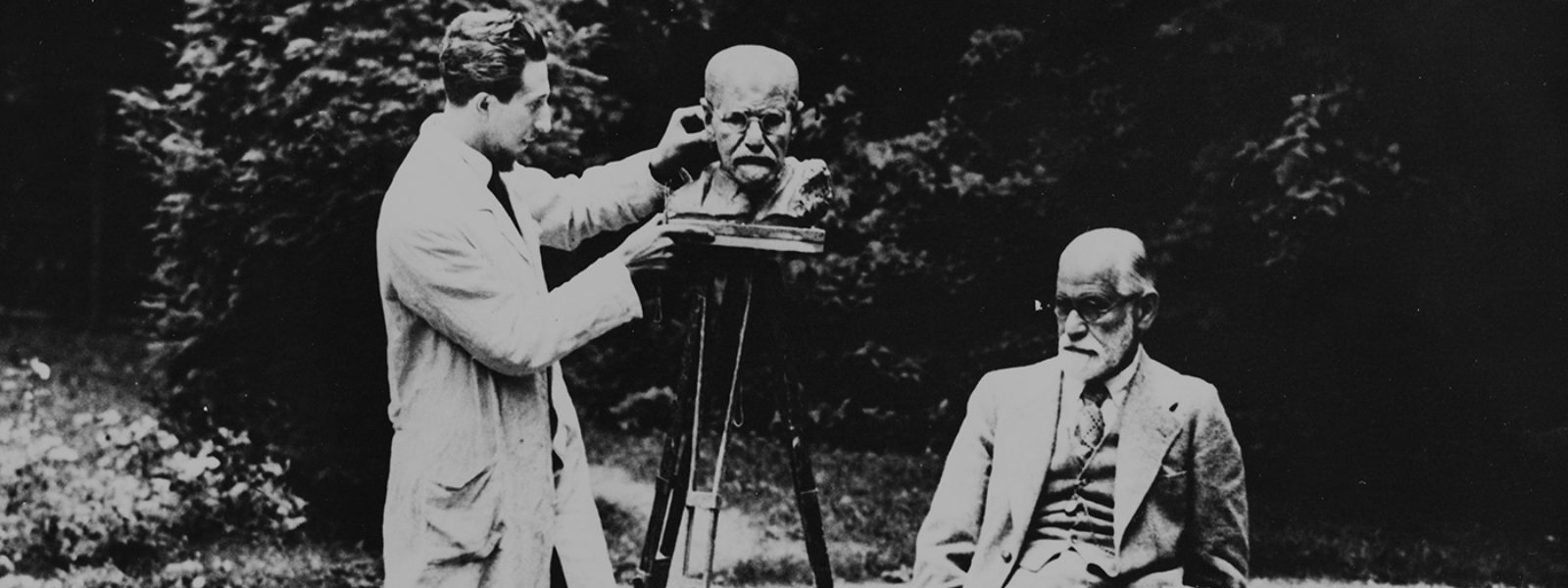 Black and white photograph of Sigmund Freud sitting in a garden while and sculptor creates a sculpture of Freud's head. A dog sits on the grass in front of them